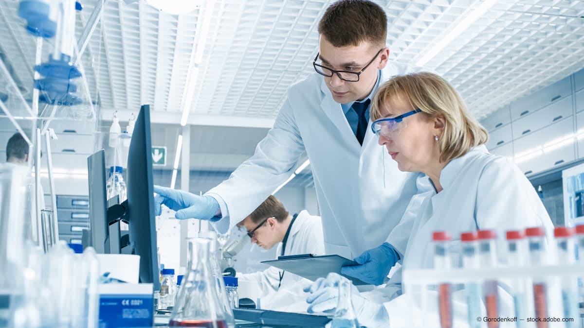 In Modern Laboratory Senior Female Scientist Has Discussion with Young Male Laboratory Assistant. He Shows Her Data Charts on a Clipboard, She Analyzes it and Enters It into Her Computer. (Adobe Stock / Gorodenkoff)